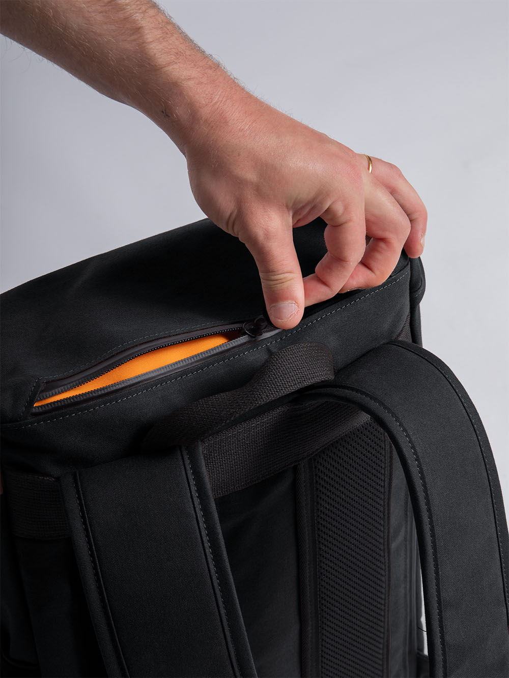 An image of a man unzipping the top quick access pocket on the Bannoch Pro - the backpack featured is in a black colour and the pocket internal colour is a bright orange. The zip is  located at the back of the backpack in the top pocket. Under nearth is the carry handle, straps and mesh back support.
