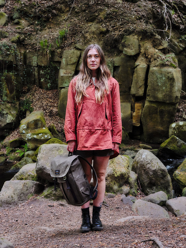 A female model in a woodland area is standing in a clearing near some rocks and a river. She is wearing a red jacket and holding the Trakke Bannoch Backpack down by her side. The Backpack is the Olive colour