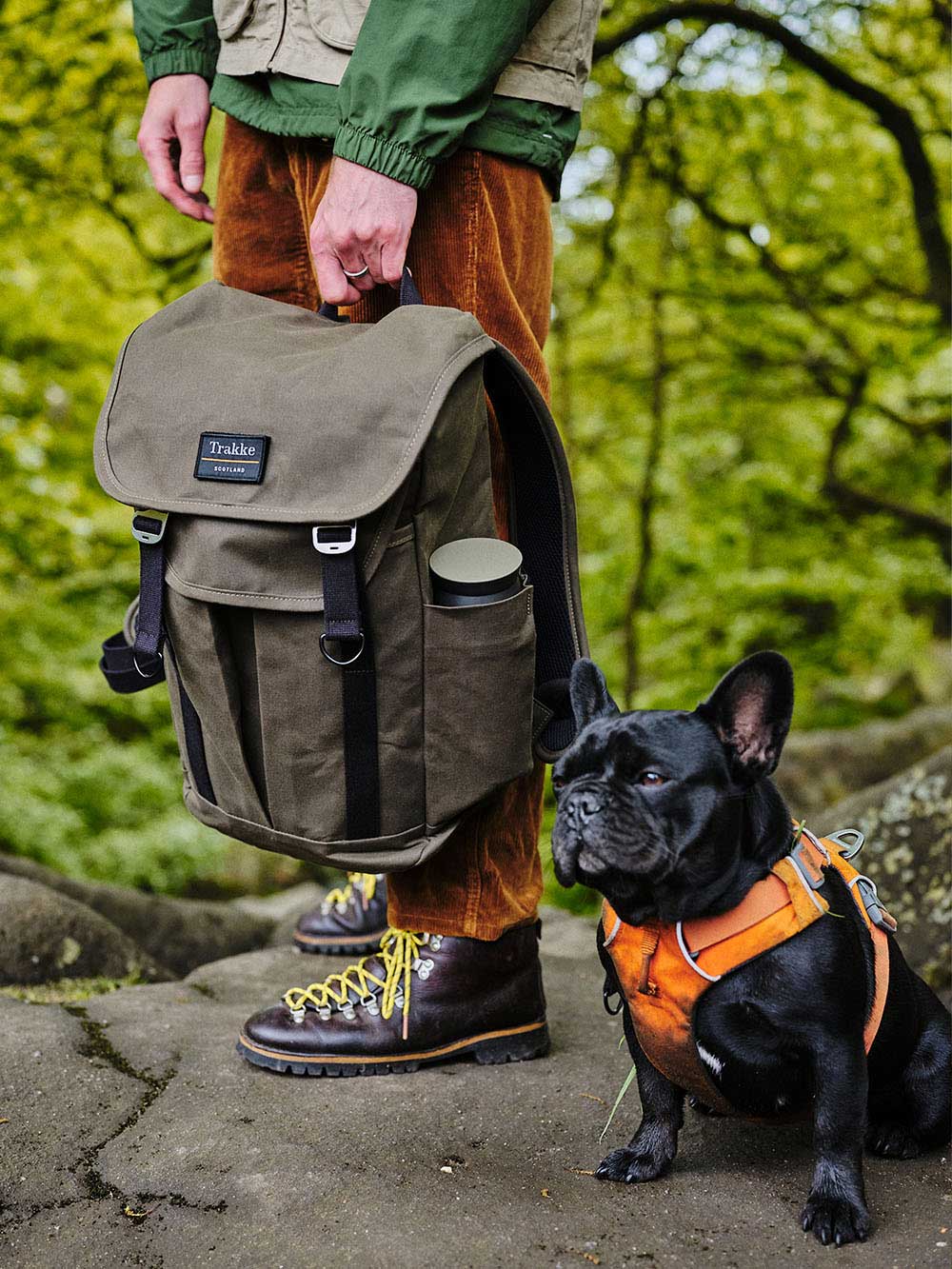 A man is holding the Trakke Bannoch in Olive against his leg, the man is standing on a rock in a woodland area outside. There is also a small Frenchie dog wearing a harness,