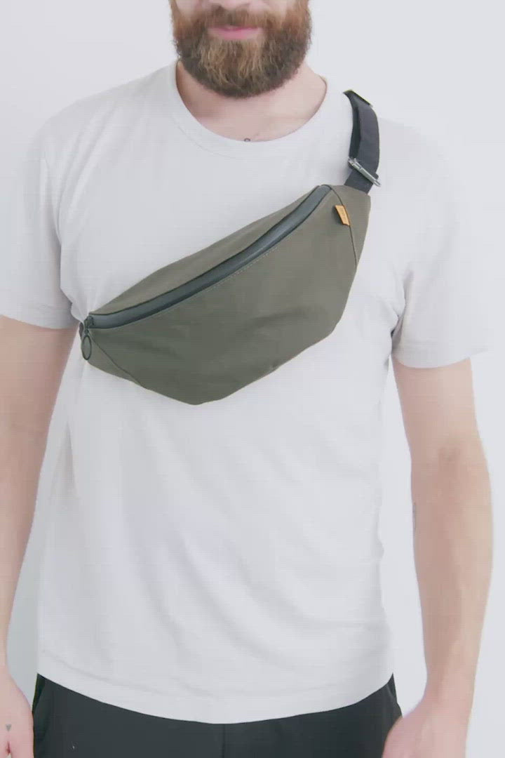 Video of man wearing a Trakke Banana Crossbody Bag in Olive. Fanny pack. Dry-finish waxed canvas. 2 litre capacity, 2L. He unzips the bag to show two internal pockets, a D-ring and orange lining.